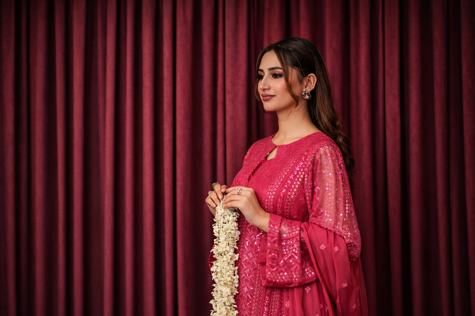 Woman in a pink traditional outfit holding a garland against a red curtain background